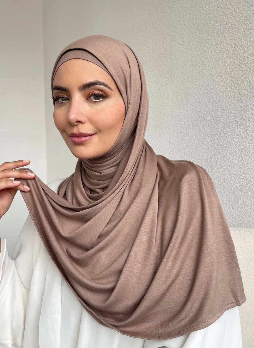 Una mujer vistiendo un hiyab beige y una blusa blanca, mirando directamente a la cámara.