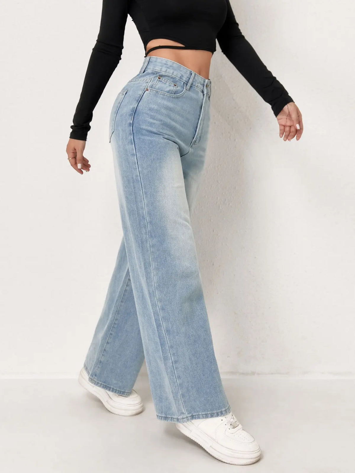 Una mujer vistiendo pantalones vaqueros anchos azul claro y una blusa negra, de pie contra una pared blanca.