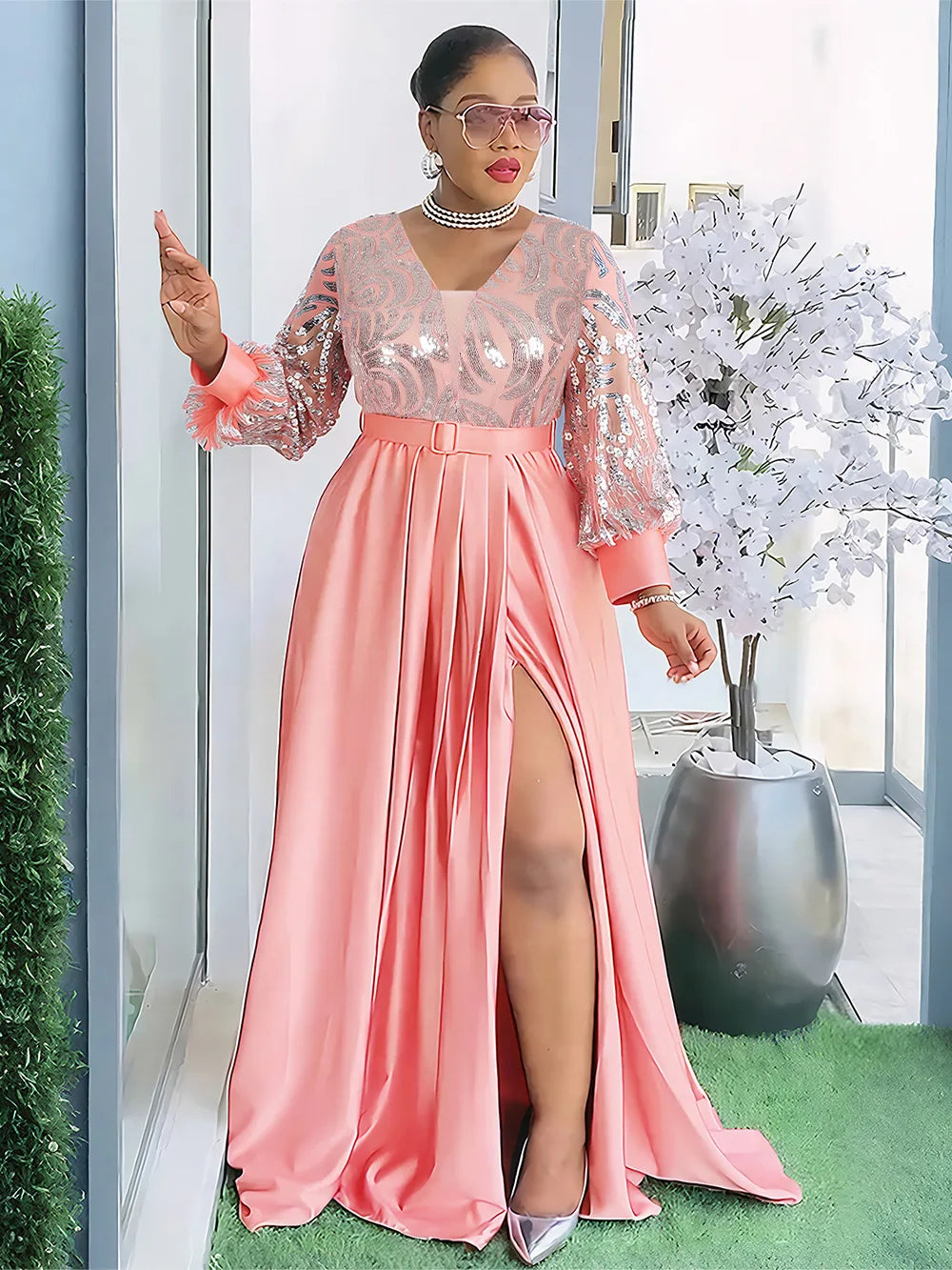 A woman models a flowing pink sequin African party dress with long sleeves and a high slit in a bright indoor setting.