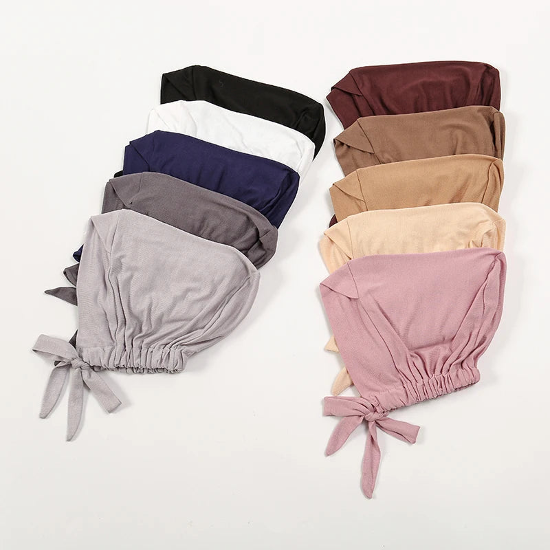A stack of various colored adjustable cotton jersey inner hijab caps in shades of black, navy, gray, beige, and pink.