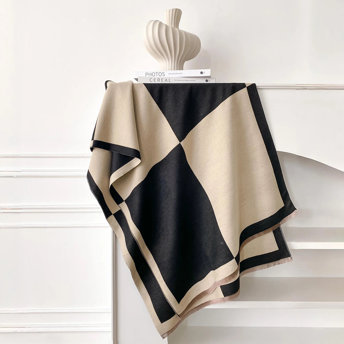 Pashmina de estampado geométrico beige y negro sobre una estantería blanca con un jarrón de cerámica.