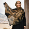 Eine Frau trägt einen braunen Tie-Dye-Hijab-Schal mit schwarzen Mustern, lächelt und blickt in die Kamera.