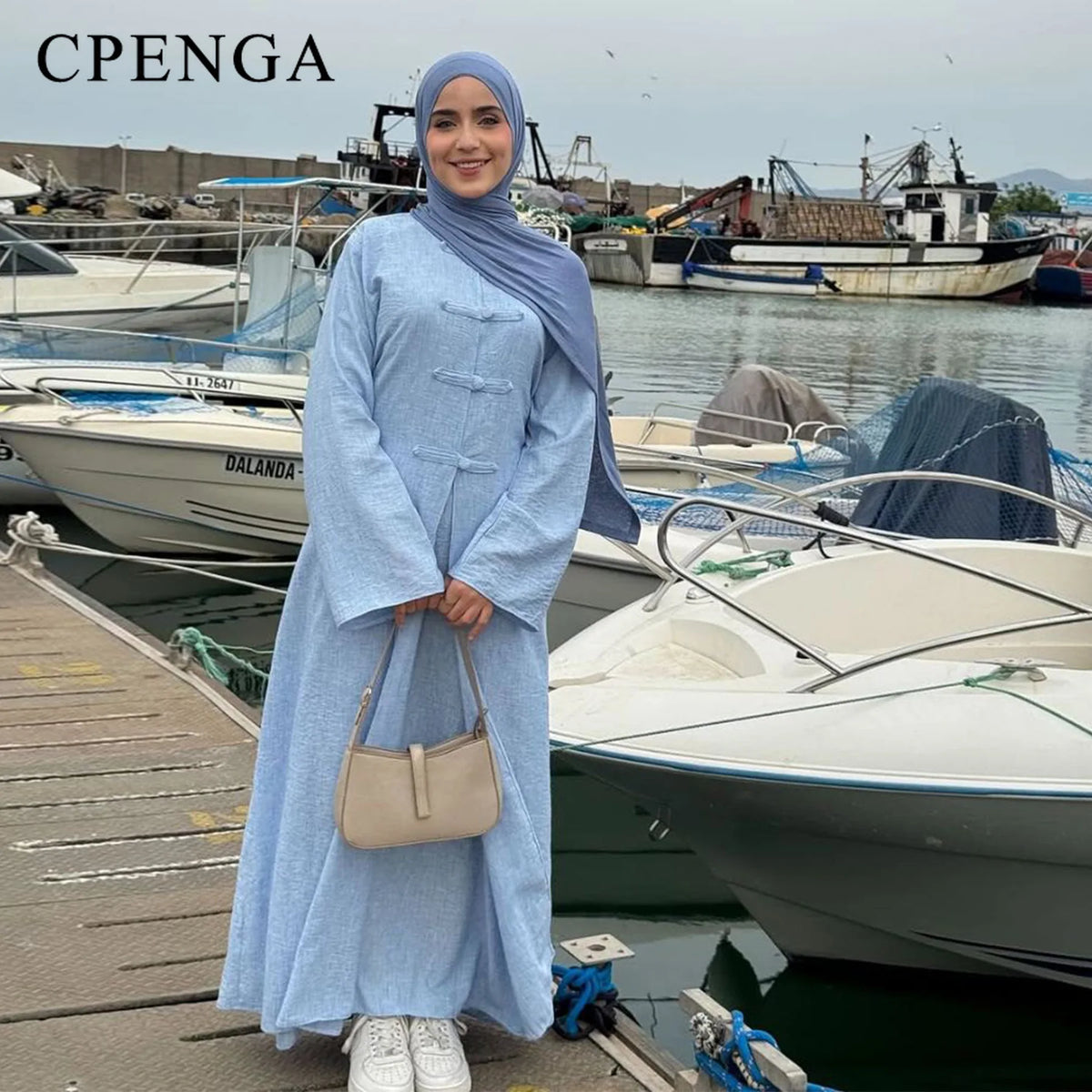 A woman wearing a light blue modest two-piece Arabic set standing on a wooden dock near boats.