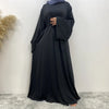 Una mujer vistiendo una elegante abaya negra con un pañuelo azul, de pie contra una pared blanca.