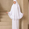 Eine Frau trägt ein weißes, mehrlagiges Spitzen-Khimar-Abaya-Set in einer neutralen Innenumgebung.