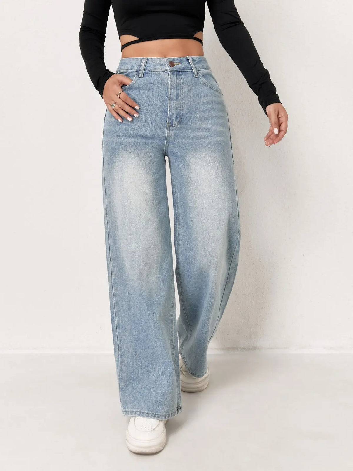 Una mujer vistiendo pantalones vaqueros anchos azul claro y una blusa negra, de pie en interiores.