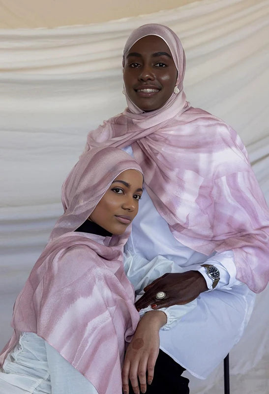 Una mujer con un pañuelo hijab estampado en acuarela rosa sostiene a otra mujer de camisa blanca.