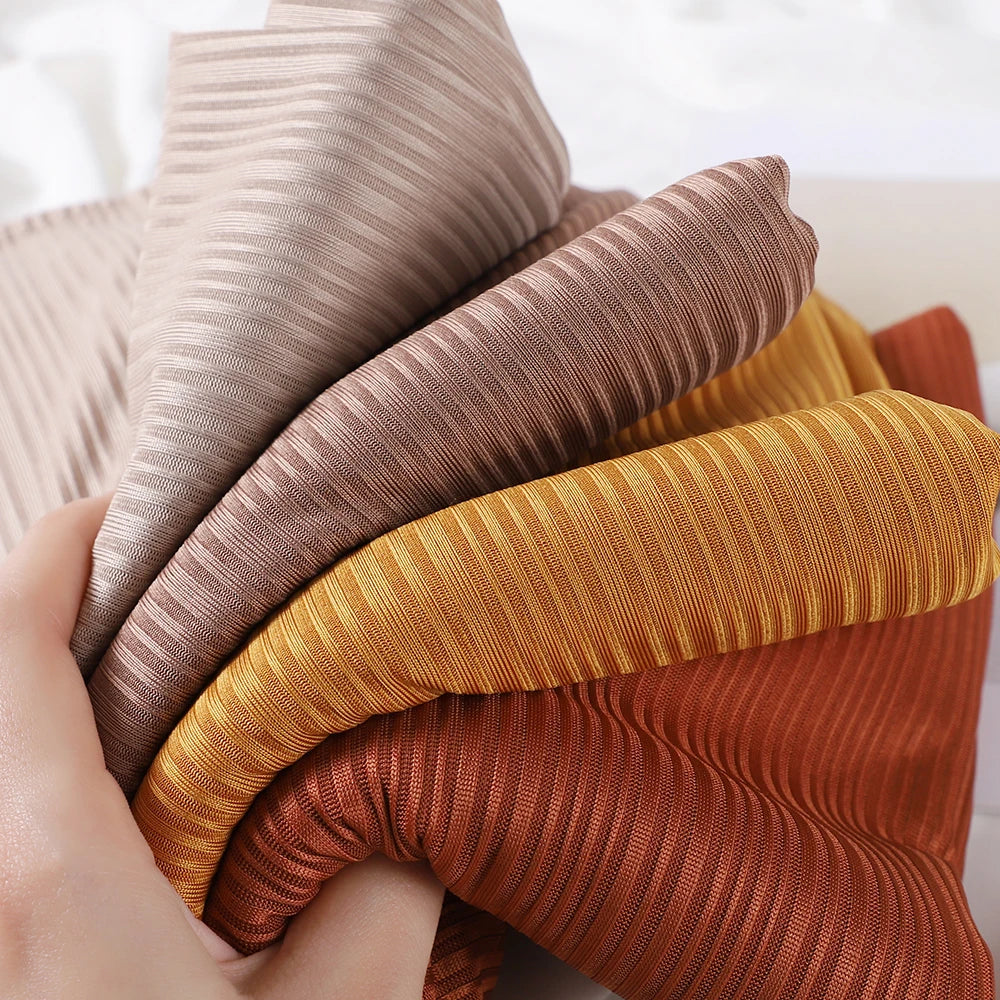 A hand is holding several rolled ribbed jersey hijab scarves in shades of beige, brown, and gold.
