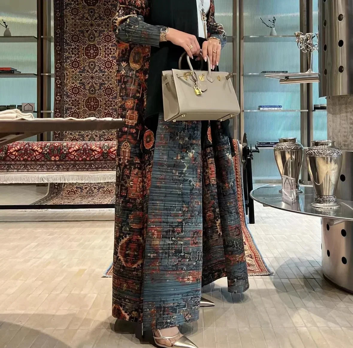 Une femme portant un long trench à motifs sur une robe noire et tenant un sac beige dans un décor moderne.