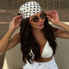 A woman wearing a white polka dot cap, sunglasses, and a white crop top and jeans.