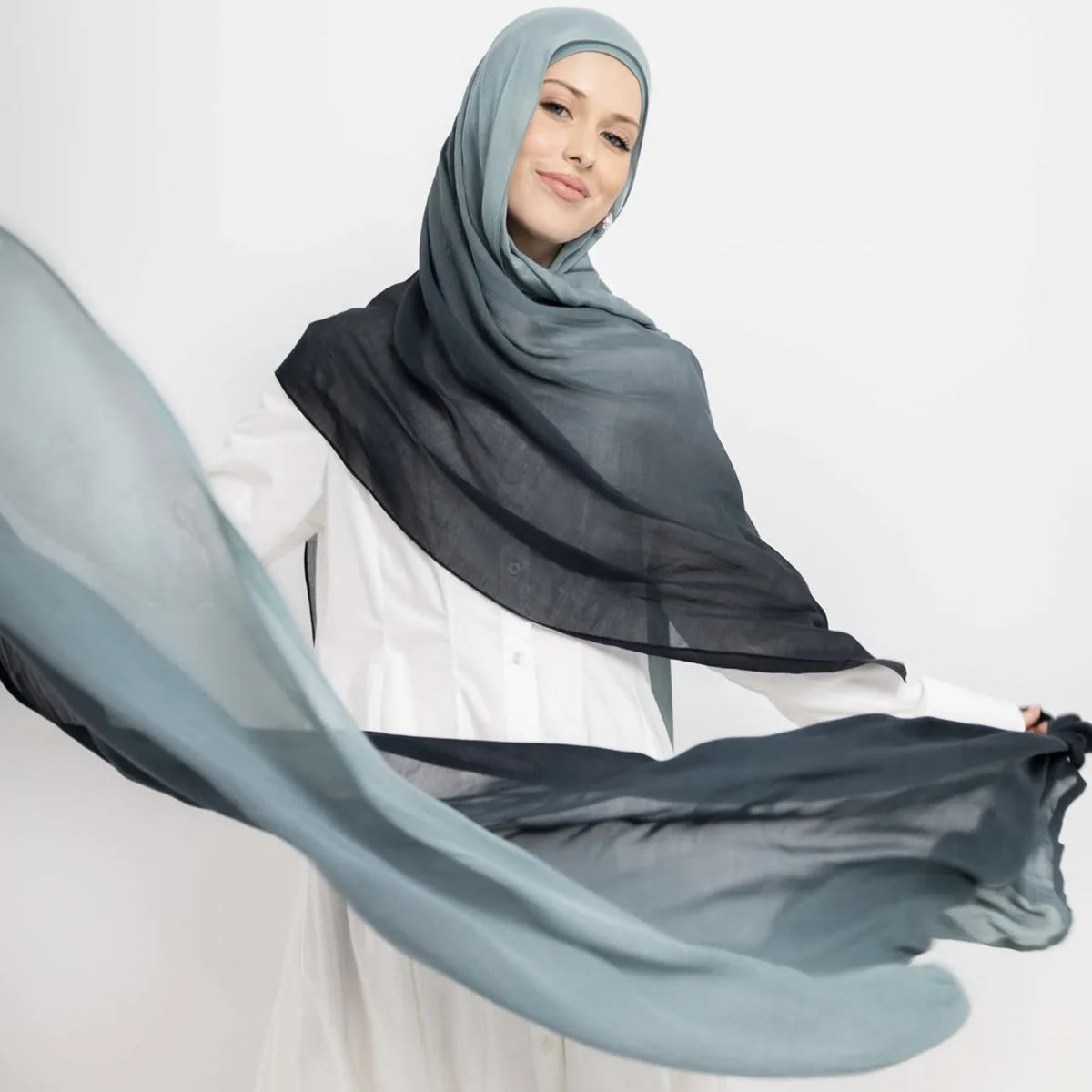 Eine Frau trägt einen grauen Ramanda Bambus Modal Rayon Schal-Hijab mit durchscheinendem blau-grauem Stoff im Vordergrund.