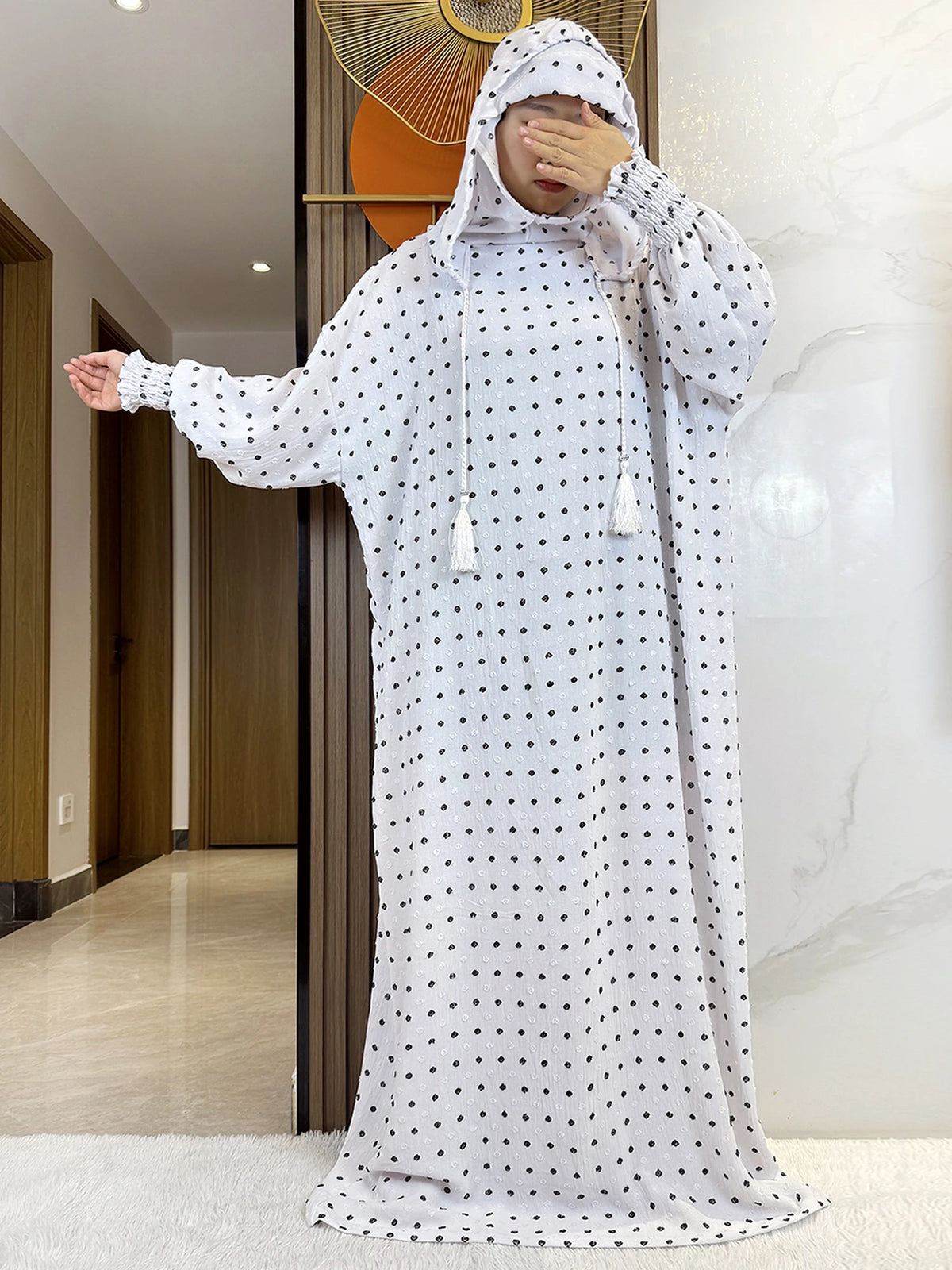 Una mujer vistiendo una abaya blanca de puntos con capucha, posando en interiores.