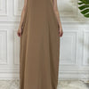 Langes, ärmelloses Kaftan-Kleid in Taupe oder Beige vor einer weißen Paneelwand.