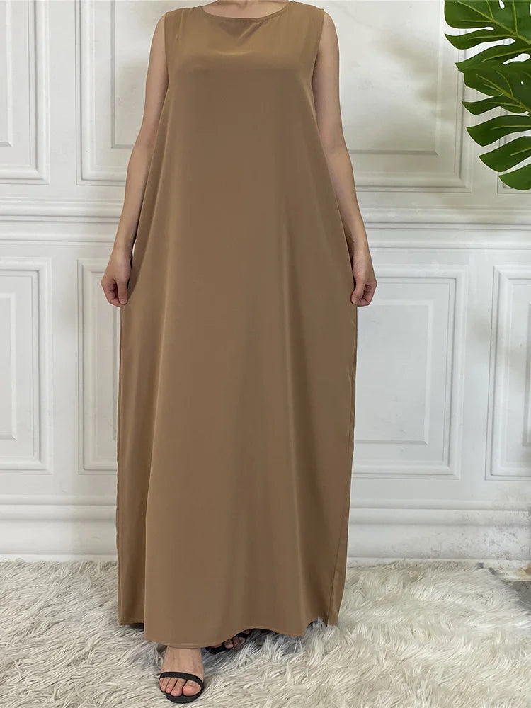 Langes, ärmelloses Kaftan-Kleid in Taupe oder Beige vor einer weißen Paneelwand.