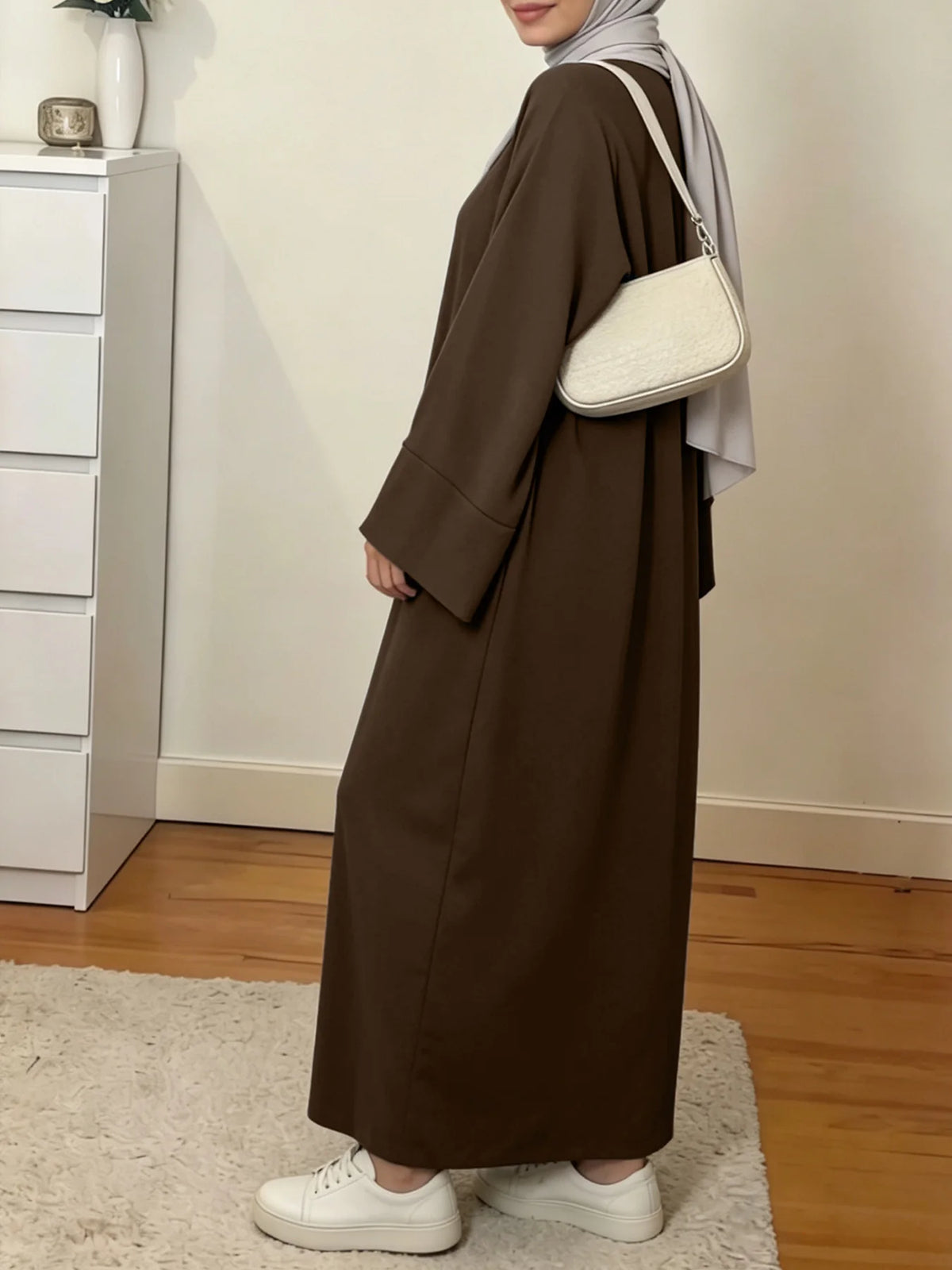 Une femme portant une élégante abaya ample marron uni et un hijab gris clair se tient dans une pièce bien éclairée.