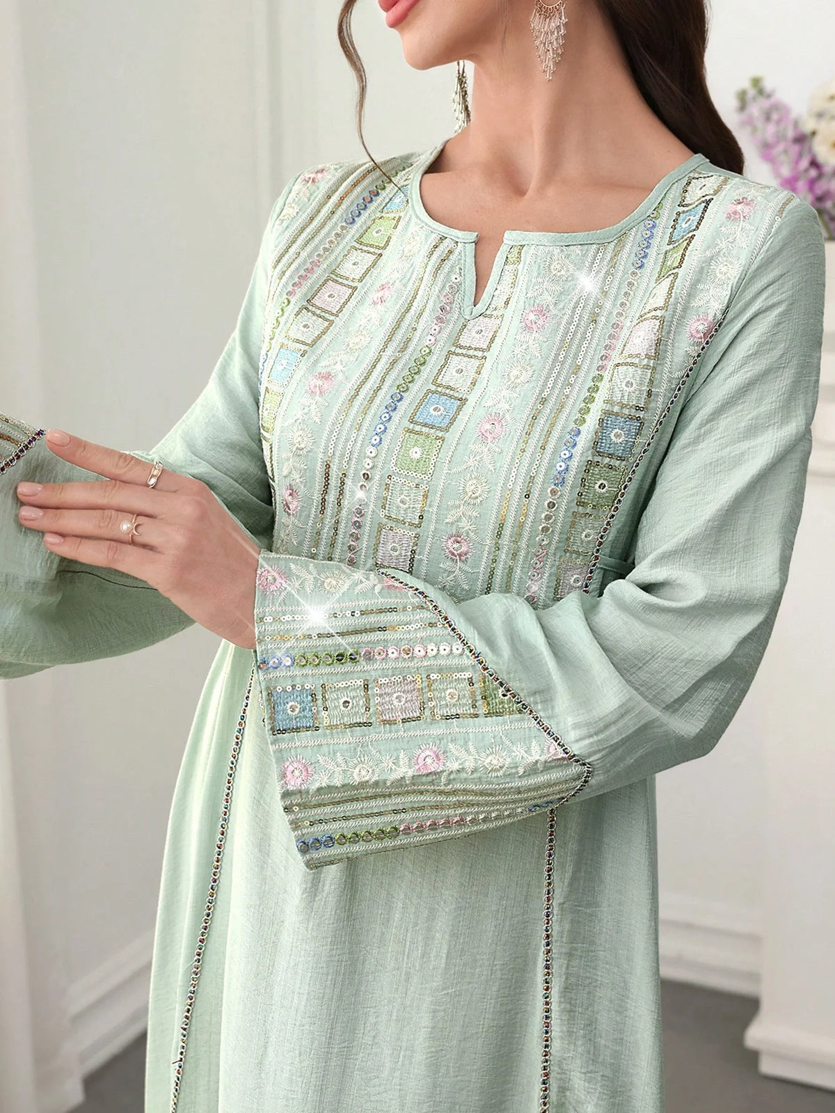 Eine Frau trägt eine hellgrüne, mit Pailletten bestickte Abaya mit zarten Blumenmustern und silbernen Ohrringen.