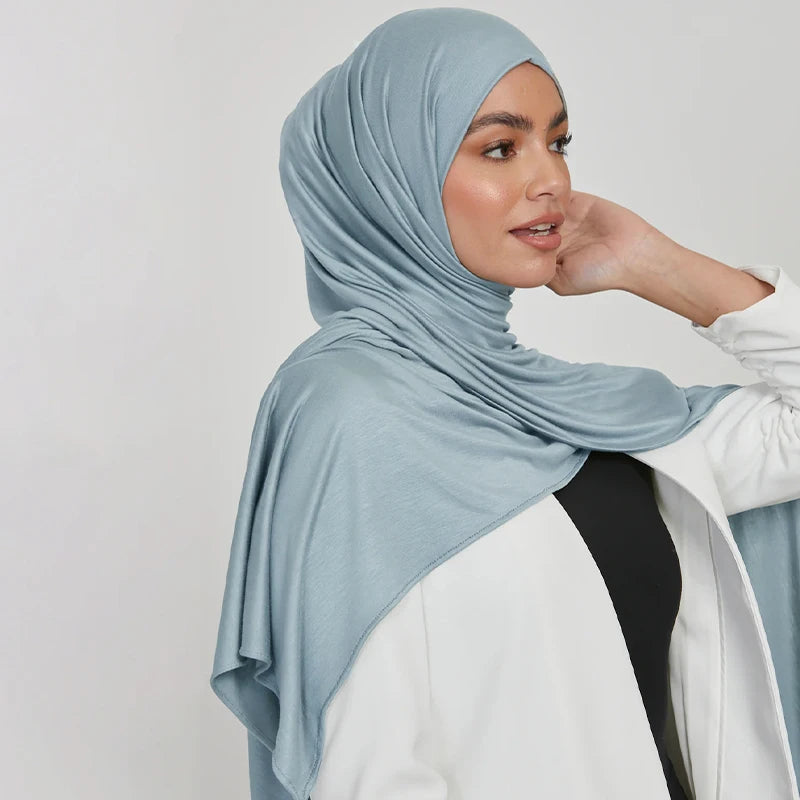 A woman wearing a light blue stretchy jersey plain hijab scarf and a white top.