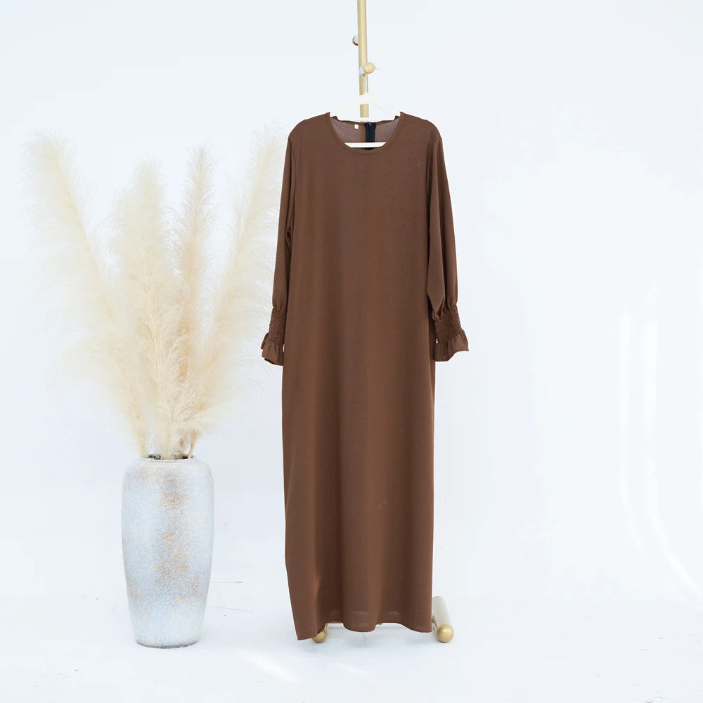Vestido abaya marrón holgado colgado en percha, junto a un jarrón de pampas grises secas.