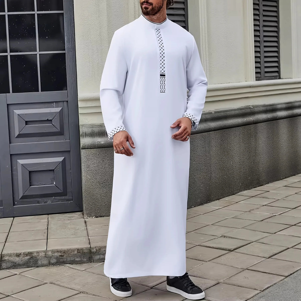 Ein Mann trägt einen weiß bestickten Jubba Kaftan mit schwarzen Akzenten, steht im Freien.