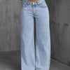 Una mujer modela pantalones vaqueros de mezclilla azul claro de pierna ancha con una blusa blanca contra un fondo gris.