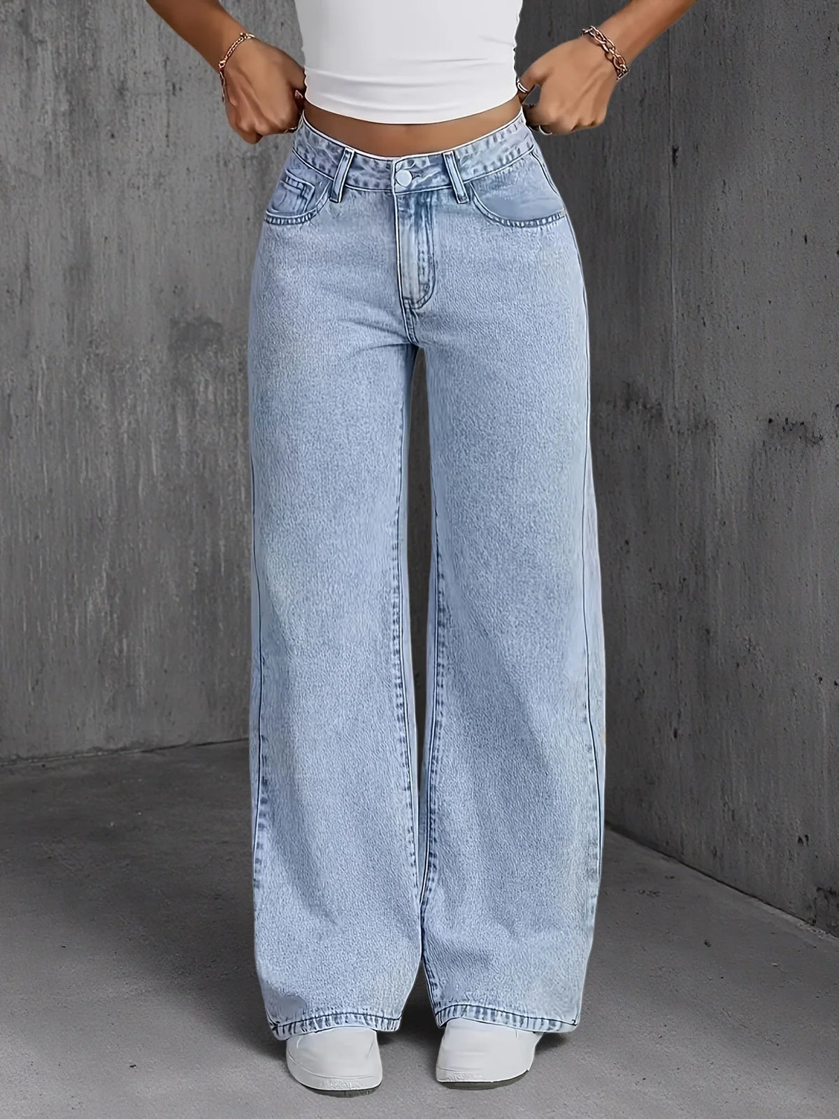 Una mujer modela pantalones vaqueros de mezclilla azul claro de pierna ancha con una blusa blanca contra un fondo gris.