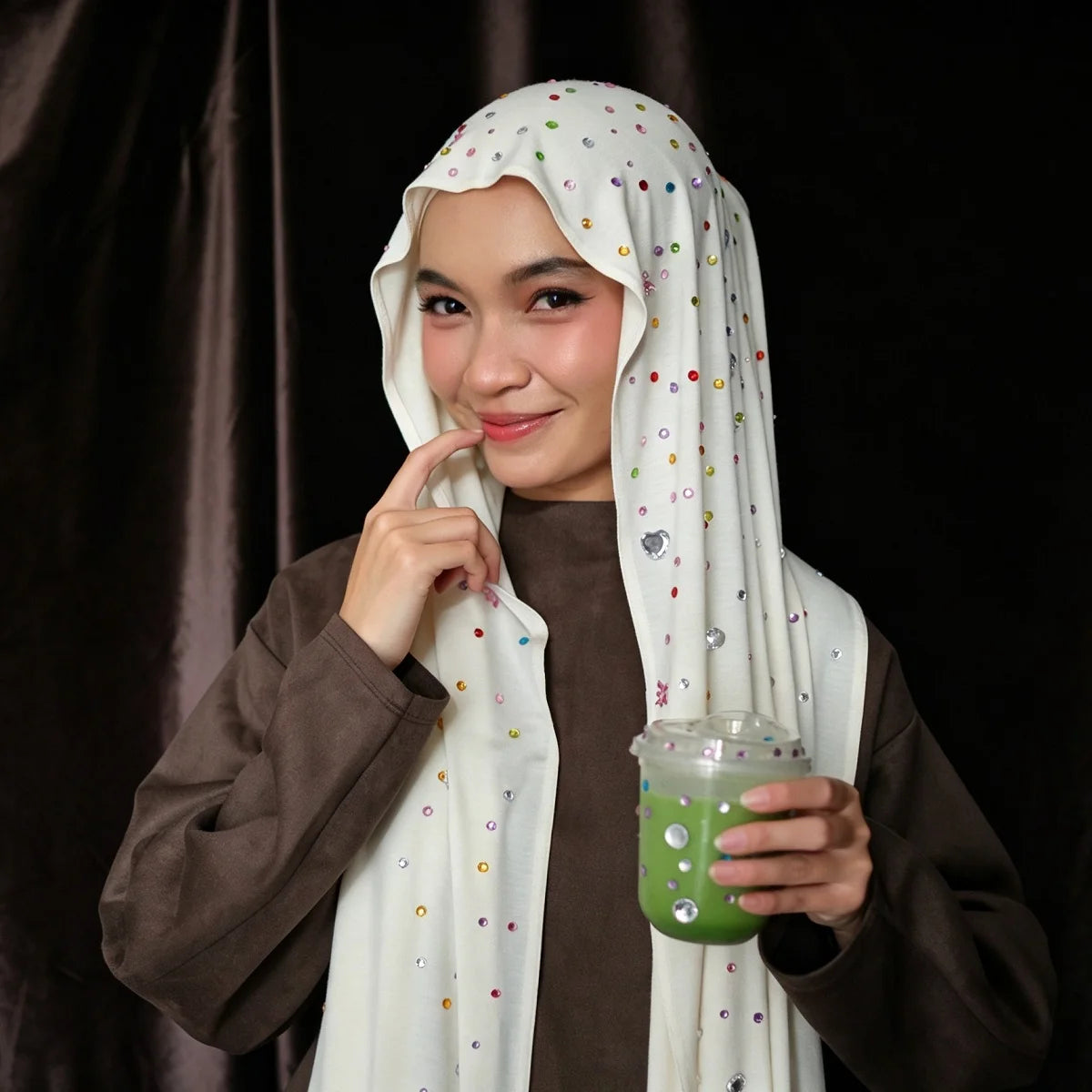 A woman wearing a white hijab scarf and brown top holds a green drink against a dark background.