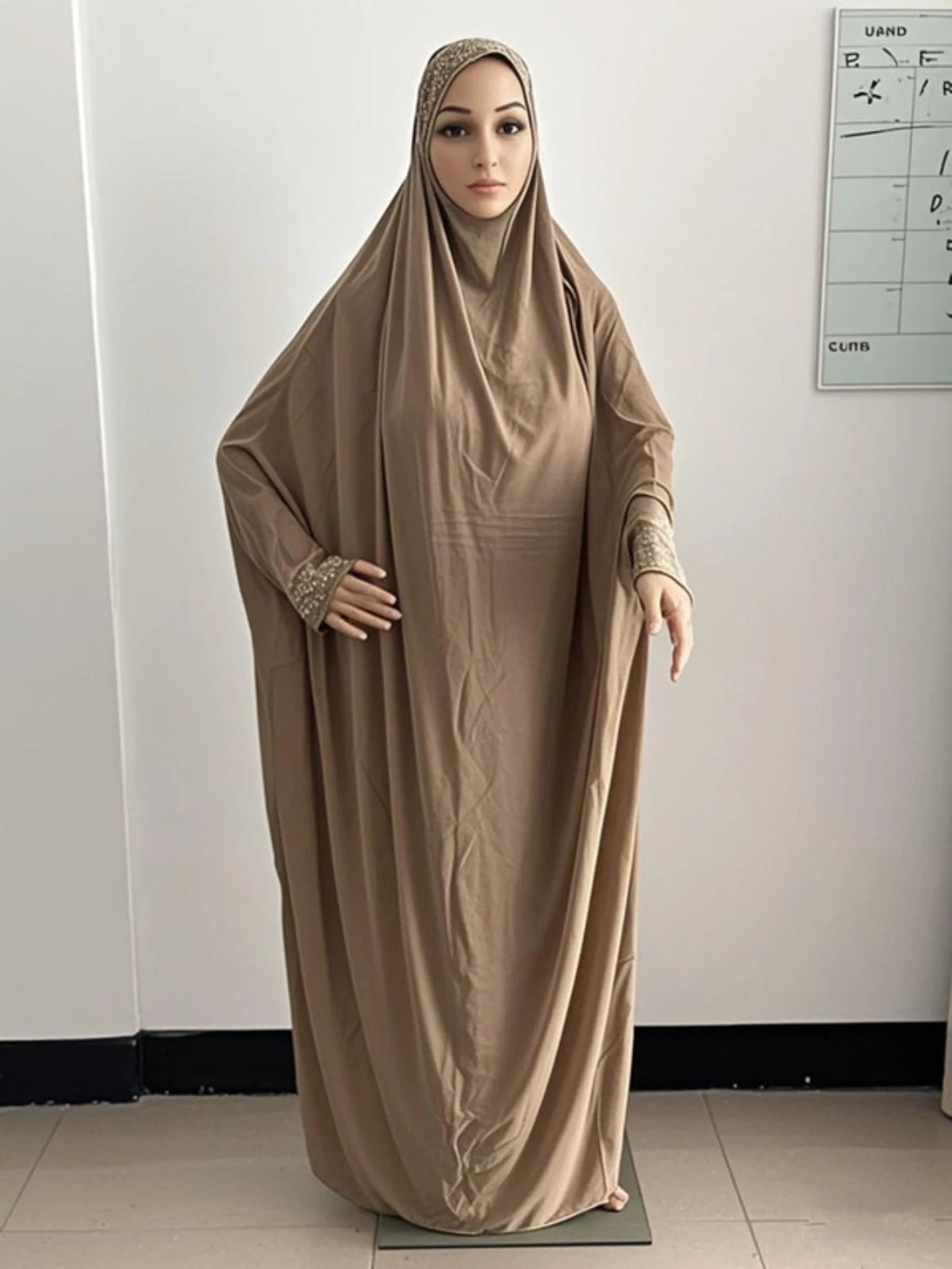 Une femme portant une abaya kaftan beige fluide avec un hijab, debout à l'intérieur.