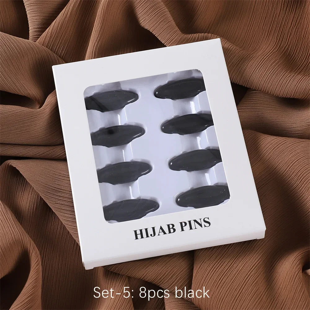 Ein Set von fünf schwarzen Hijab-Pins in einer weißen Box auf einem braunen Stoffhintergrund.
