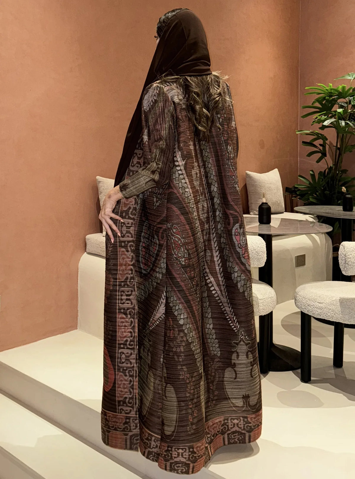 Una mujer vistiendo un cárdigan abaya plisado marrón oscuro con hijab, exhibido en un pedestal blanco.