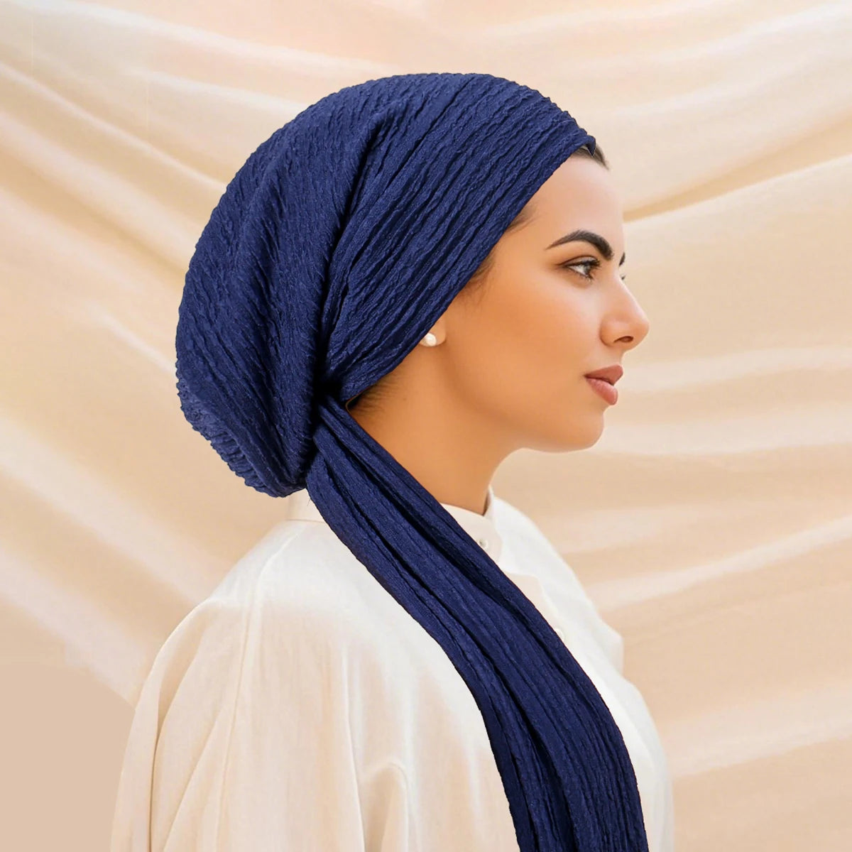 A woman wearing a dark blue solid color turban hijab cap and a cream-colored top.