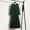 Dunkelgrünes Maxi-Kleid mit langen Ärmeln an einem schwarzen Haken vor einer weißen Wand.