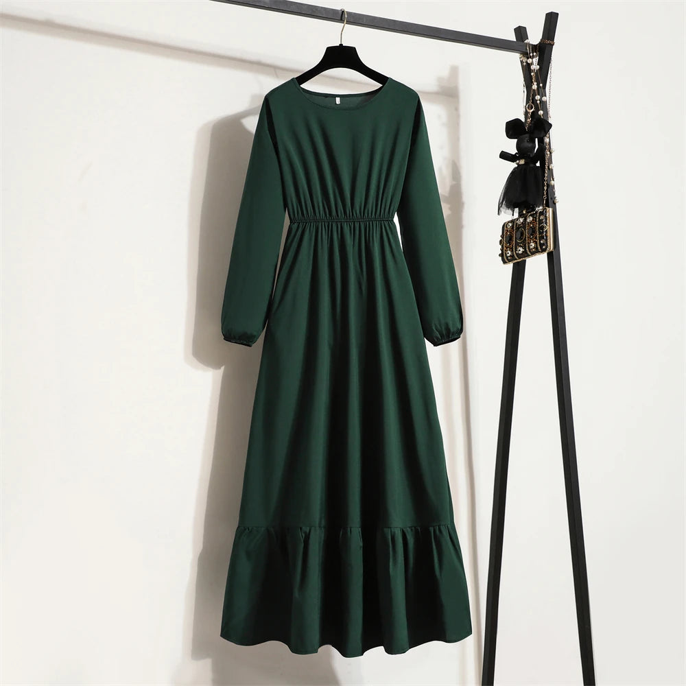 Dunkelgrünes Maxi-Kleid mit langen Ärmeln an einem schwarzen Haken vor einer weißen Wand.