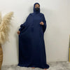 Une femme portant une abaya bleu foncé avec capuche et manches batwing, debout contre un mur blanc.
