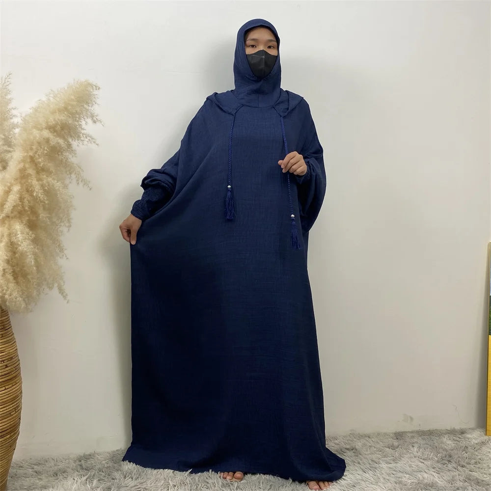 Une femme portant une abaya bleu foncé avec capuche et manches batwing, debout contre un mur blanc.