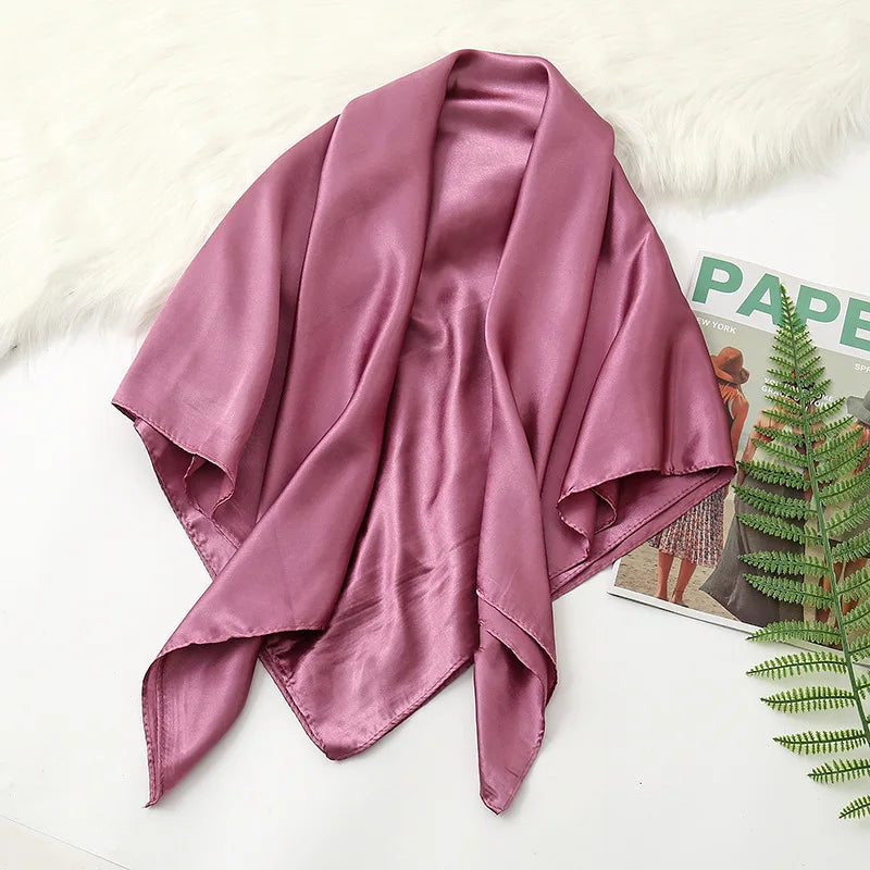 A draped, solid mauve silk satin hijab scarf laid on a white surface next to a green fern.