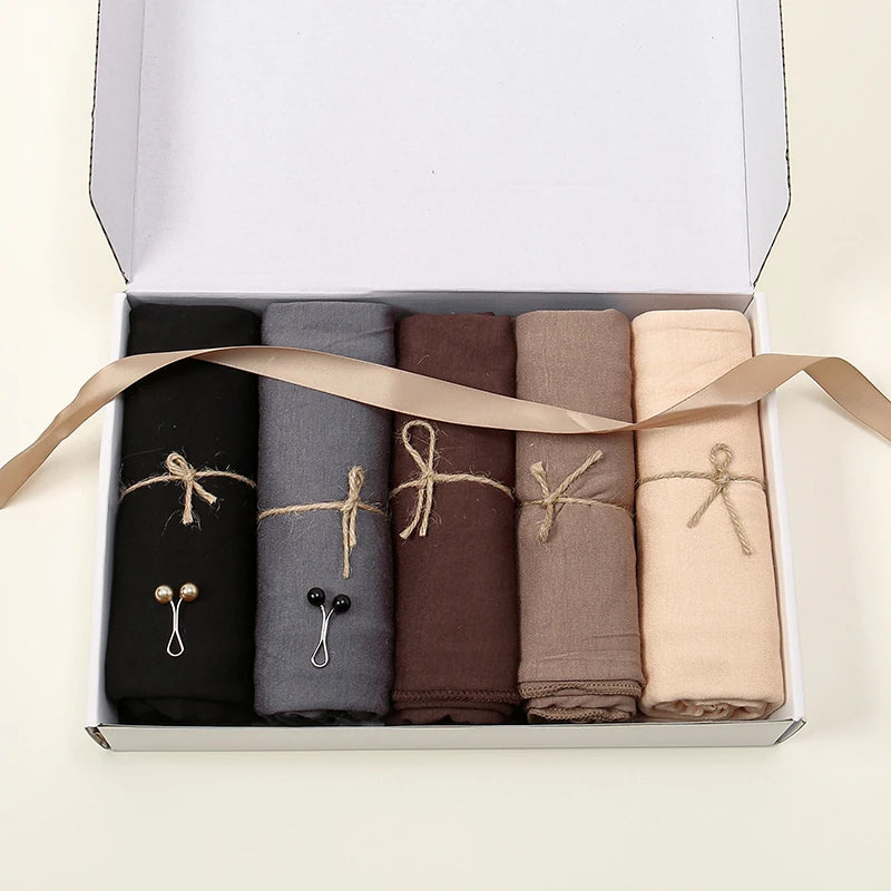 Une boîte contenant cinq foulards hijab solides roulés de couleur noire, grise, marron, beige et crème.