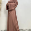 Eine Frau trägt ein fließendes, offenes Abaya-Kaftan in staubrosa Farbe und einen weißen Hijab.