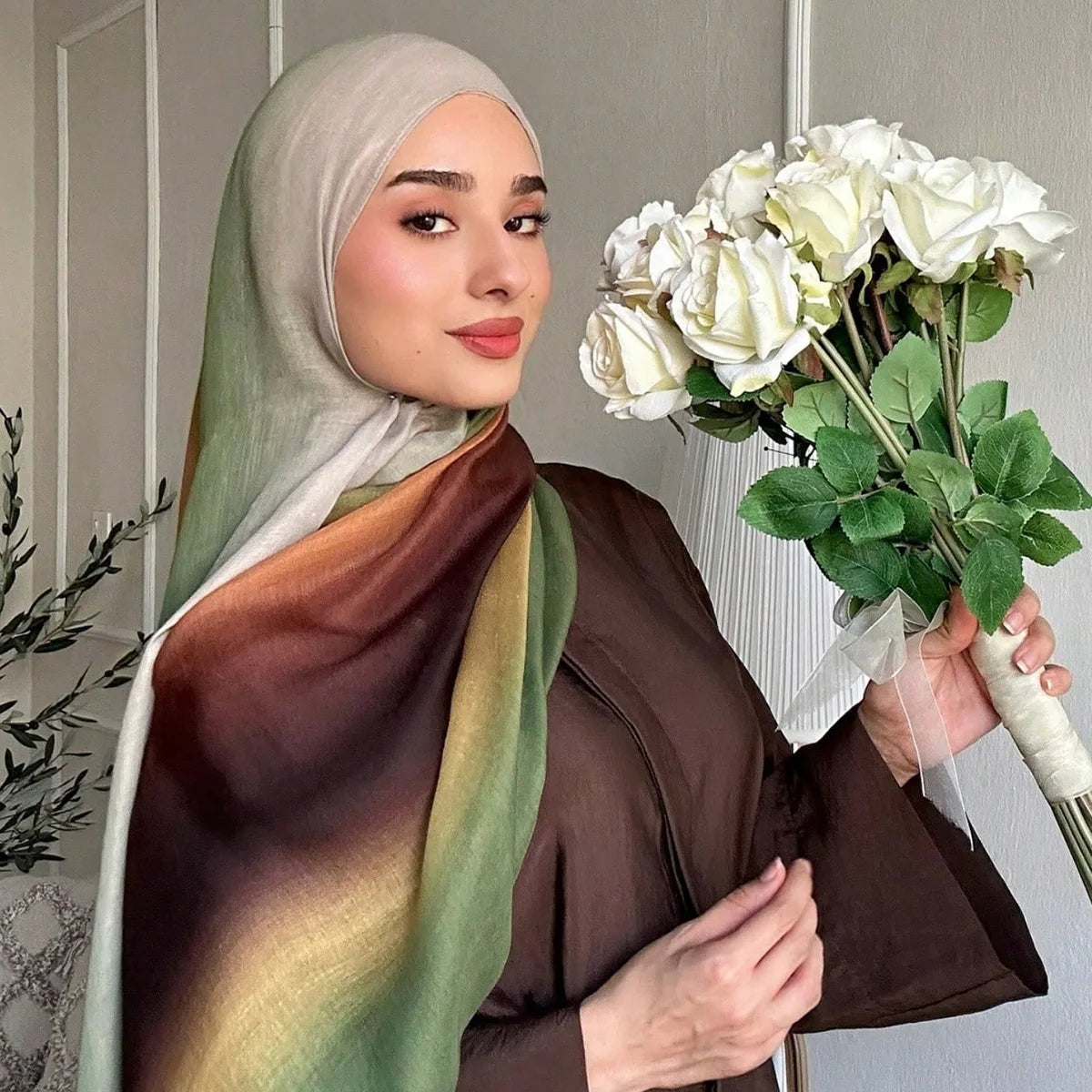 Eine Frau trägt einen Leinen-Hijab-Schal mit Aquarelldruck in Olivgrün- und Brauntönen und hält einen Strauß weißer Rosen