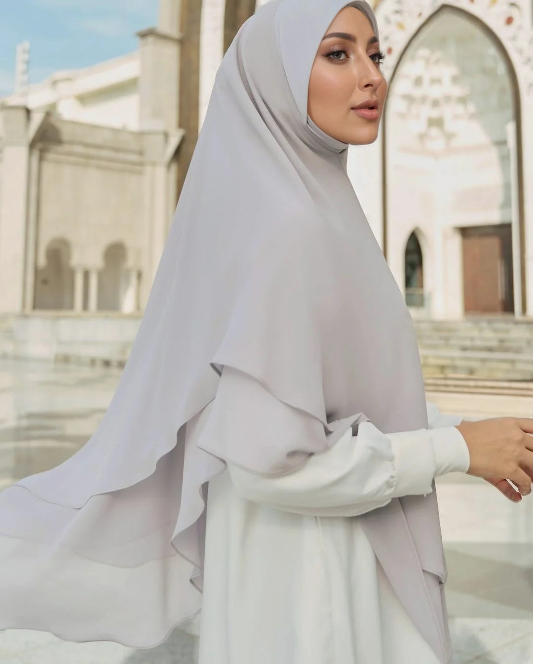 Eine Frau trägt einen hellgrauen doppelten Chiffon-Instant-Hijab und weiße Kleidung in einer Außenarchitekturumgebung.