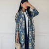 Una mujer vistiendo una chaqueta abaya de color azul con estampado floral, capa interior negra y pantalones blancos.