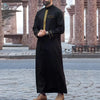 A man wearing a black Moroccan embroidered thobe jellaba standing in an outdoor courtyard.