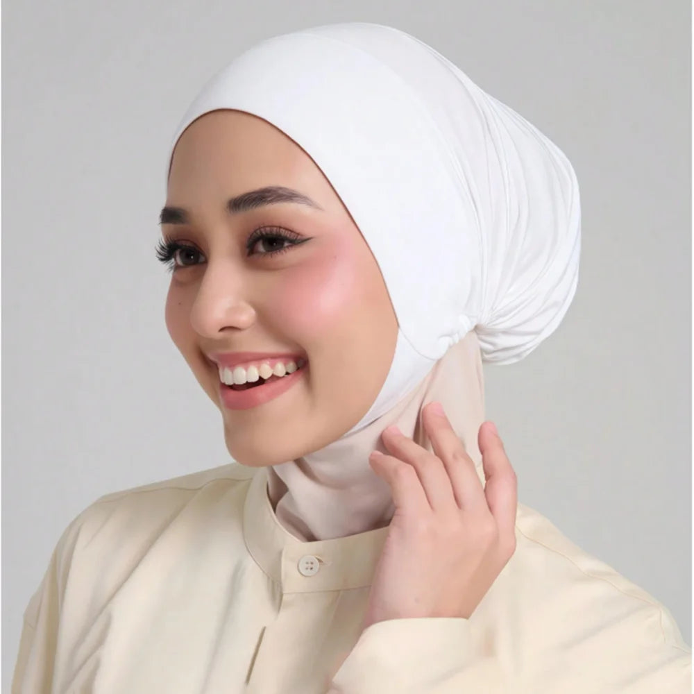Une femme portant un hijab blanc uni et un haut de couleur crème, souriante et touchant son cou.