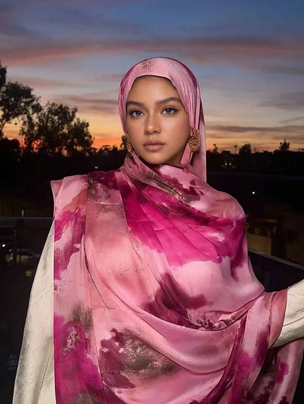 A woman wearing a pink and purple shimmer satin silk hijab shawl against an orange sunset background.