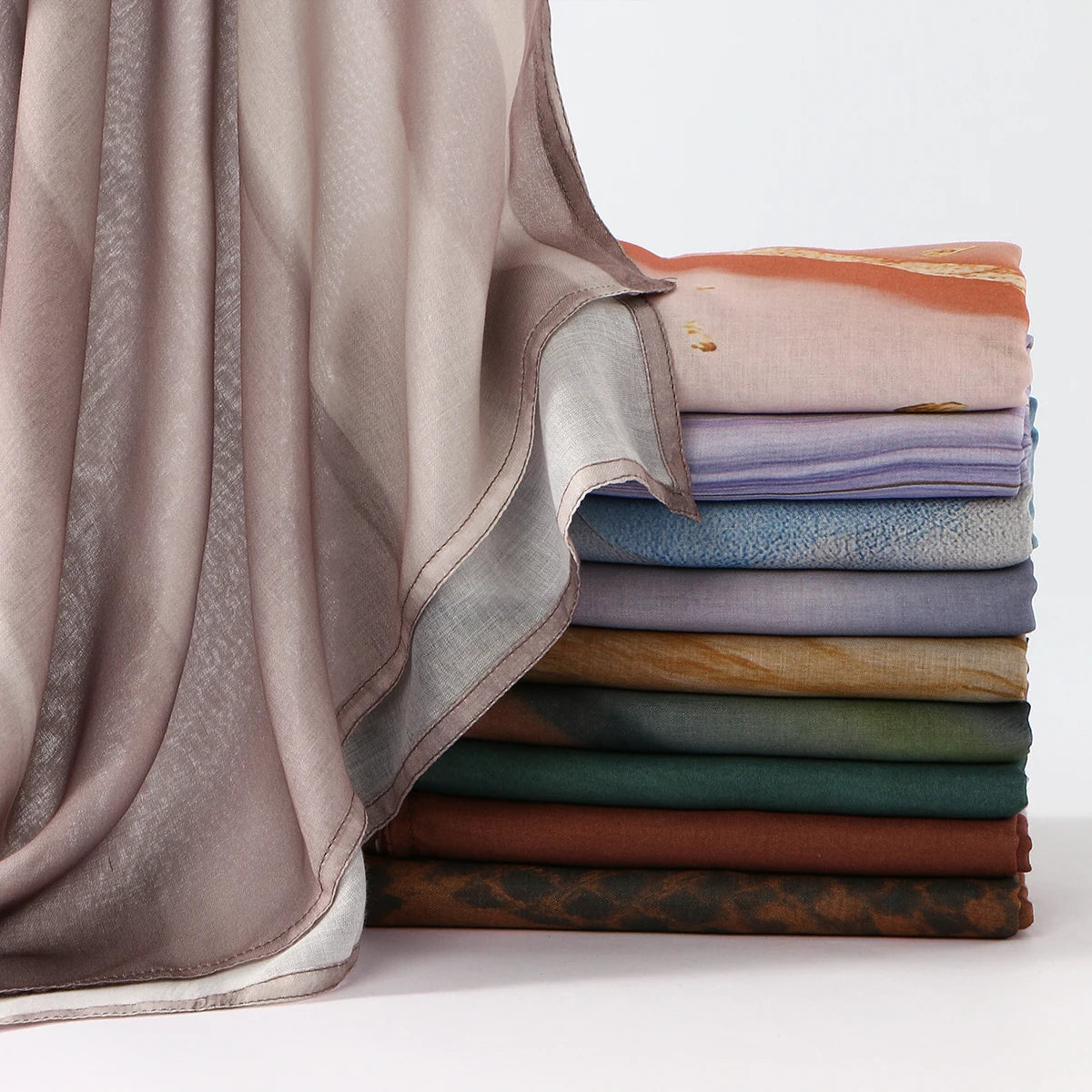 A stack of elegant printed linen hijab scarves in various muted tones, including beige, purple, and olive green.