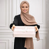 Une femme portant un hijab beige tient une boîte cadeau rose avec broderie florale.