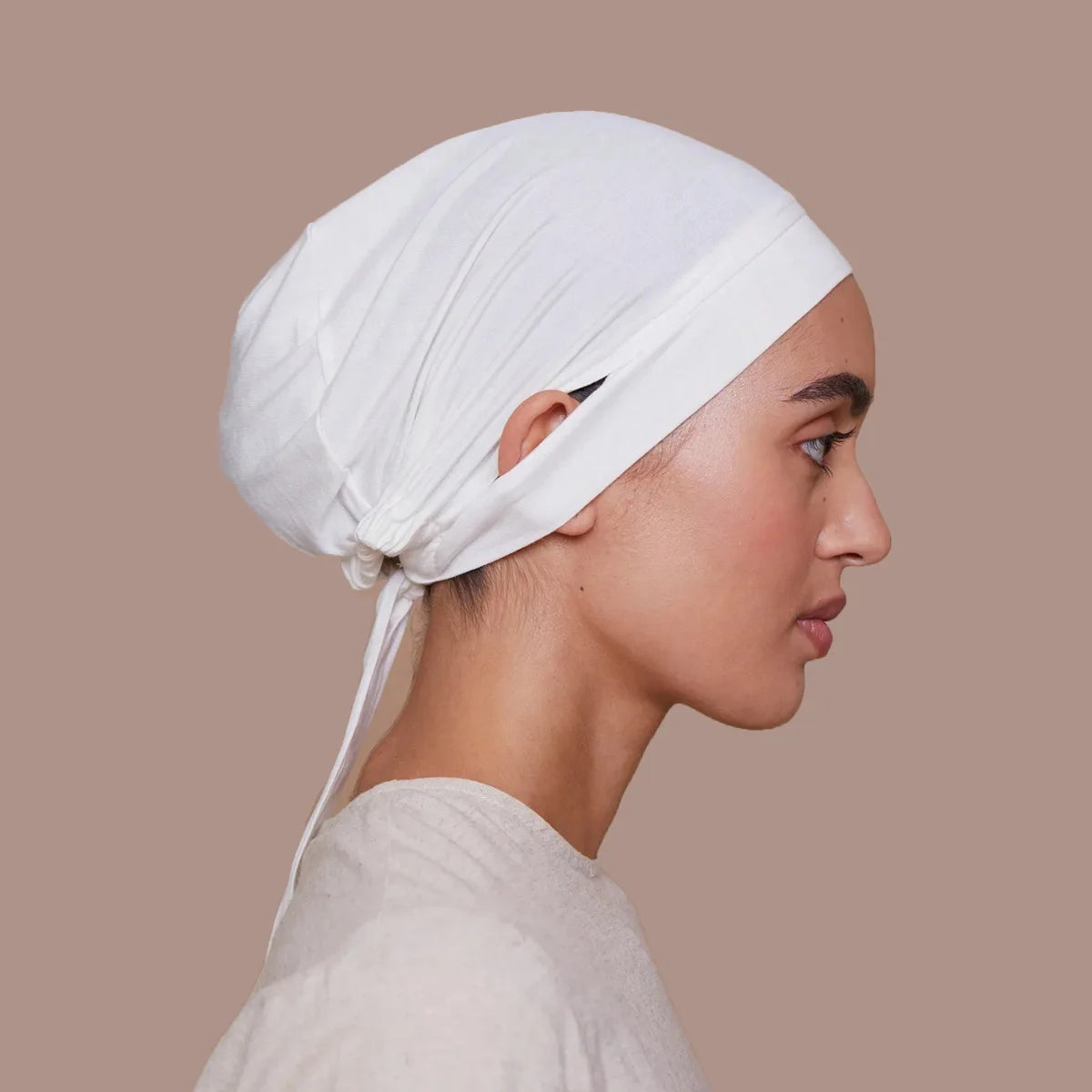 Une femme portant un bonnet de hijab intérieur blanc à cordon, vue de profil sur fond neutre.