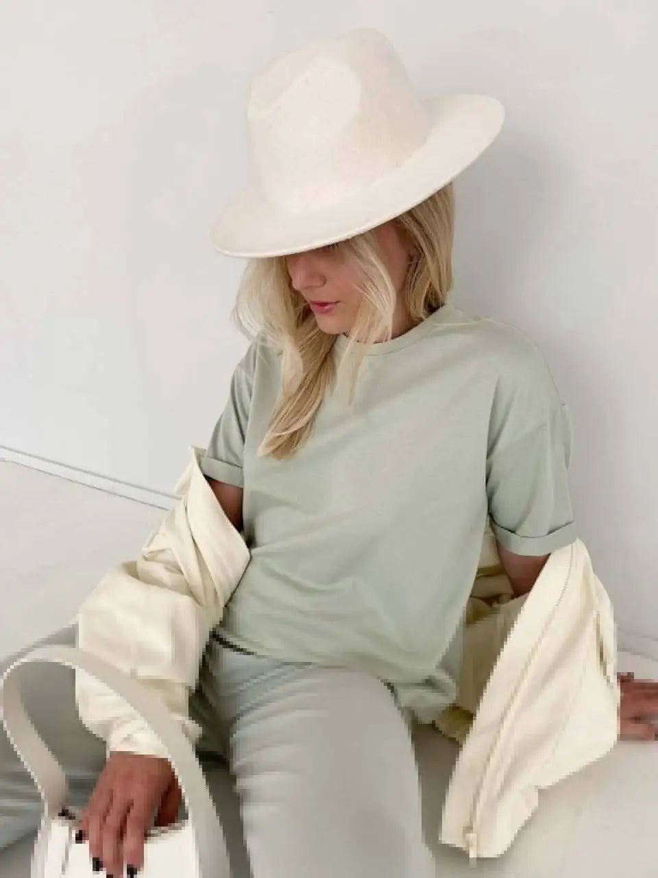 Una mujer rubia vistiendo una camiseta básica de algodón verde claro oversize y pantalones blancos, sosteniendo una chaqueta 