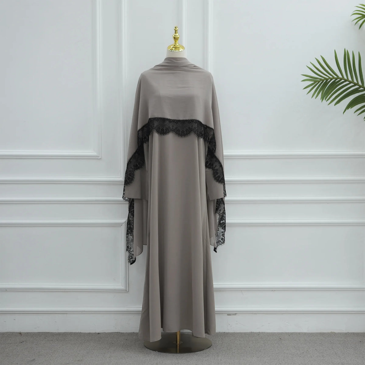 Vestido abaya largo color topo con ribetes de encaje negro y un broche dorado, exhibido en maniquí.