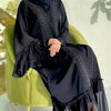 Eine Frau trägt eine elegante schwarze, bodenlange Abaya mit Spitzenverzierungen und sitzt auf einem grünen Stuhl.
