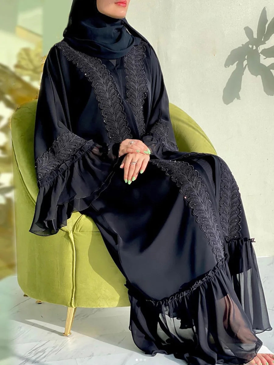 Eine Frau trägt eine elegante schwarze, bodenlange Abaya mit Spitzenverzierungen und sitzt auf einem grünen Stuhl.